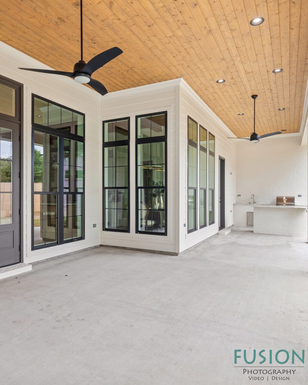 A covered outdoor patio featuring a concrete floor, a wood-paneled ceiling with two black ceiling fans, white walls, black-framed windows, and a built-in outdoor grill area.