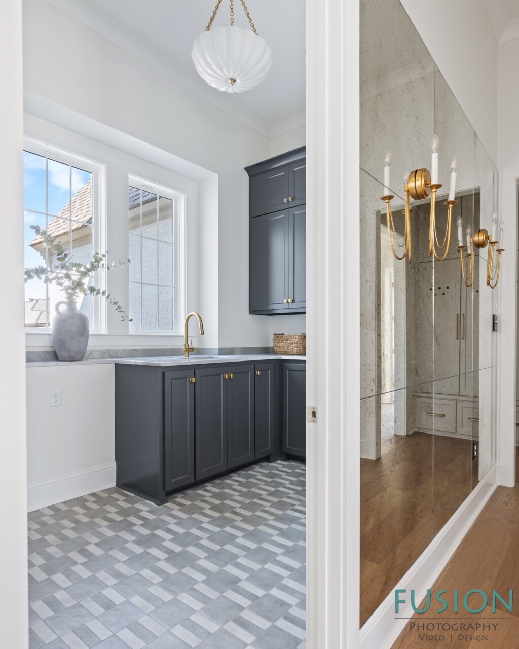 A kitchen or utility space with dark grey cabinets, a white countertop, and a checkered floor, viewed through a doorway beside a wall of antiqued mirrors.