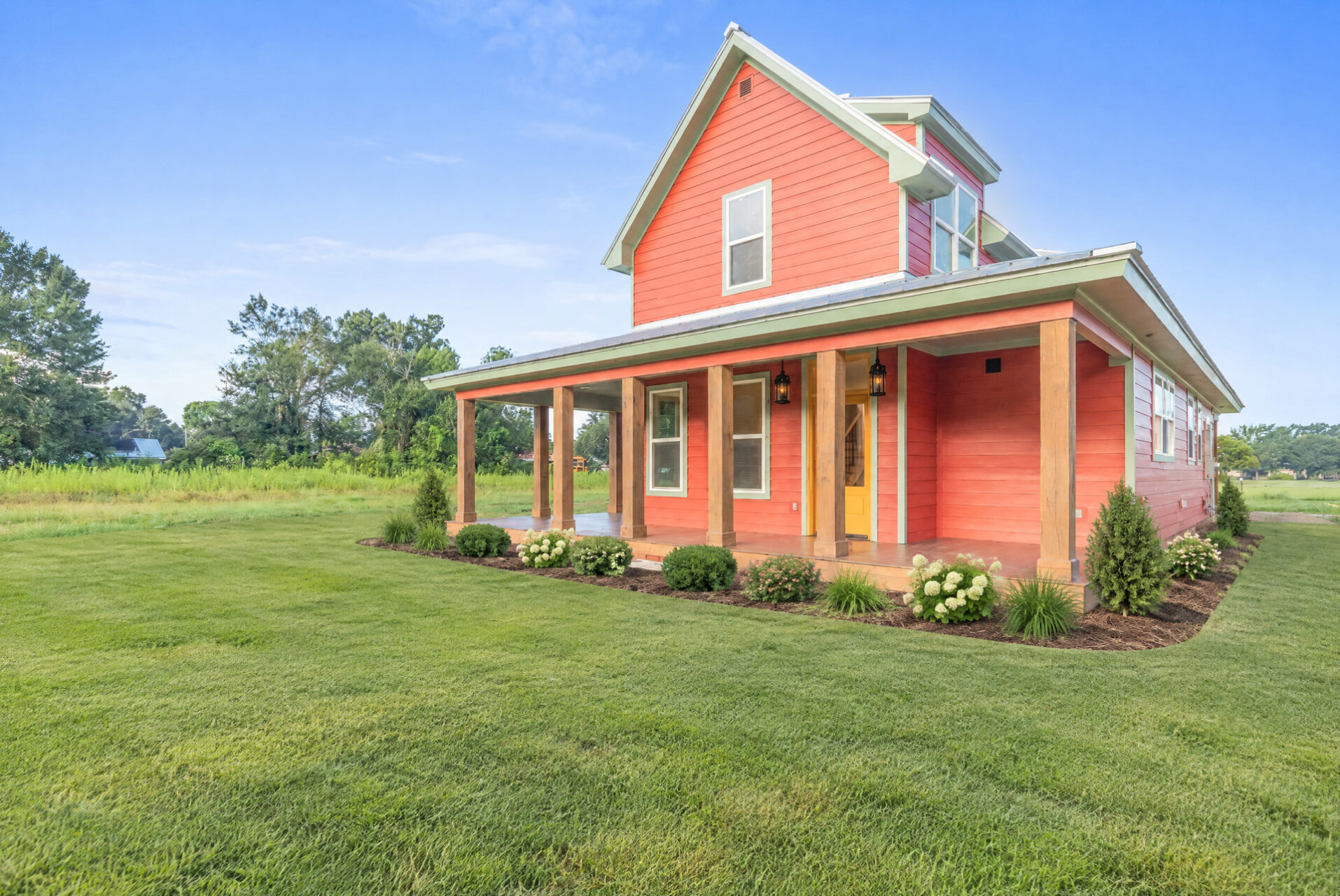 A two-story coral-colored house with a wrap-around porch, wooden support beams, and a bright yellow front door, situated in a grassy field.