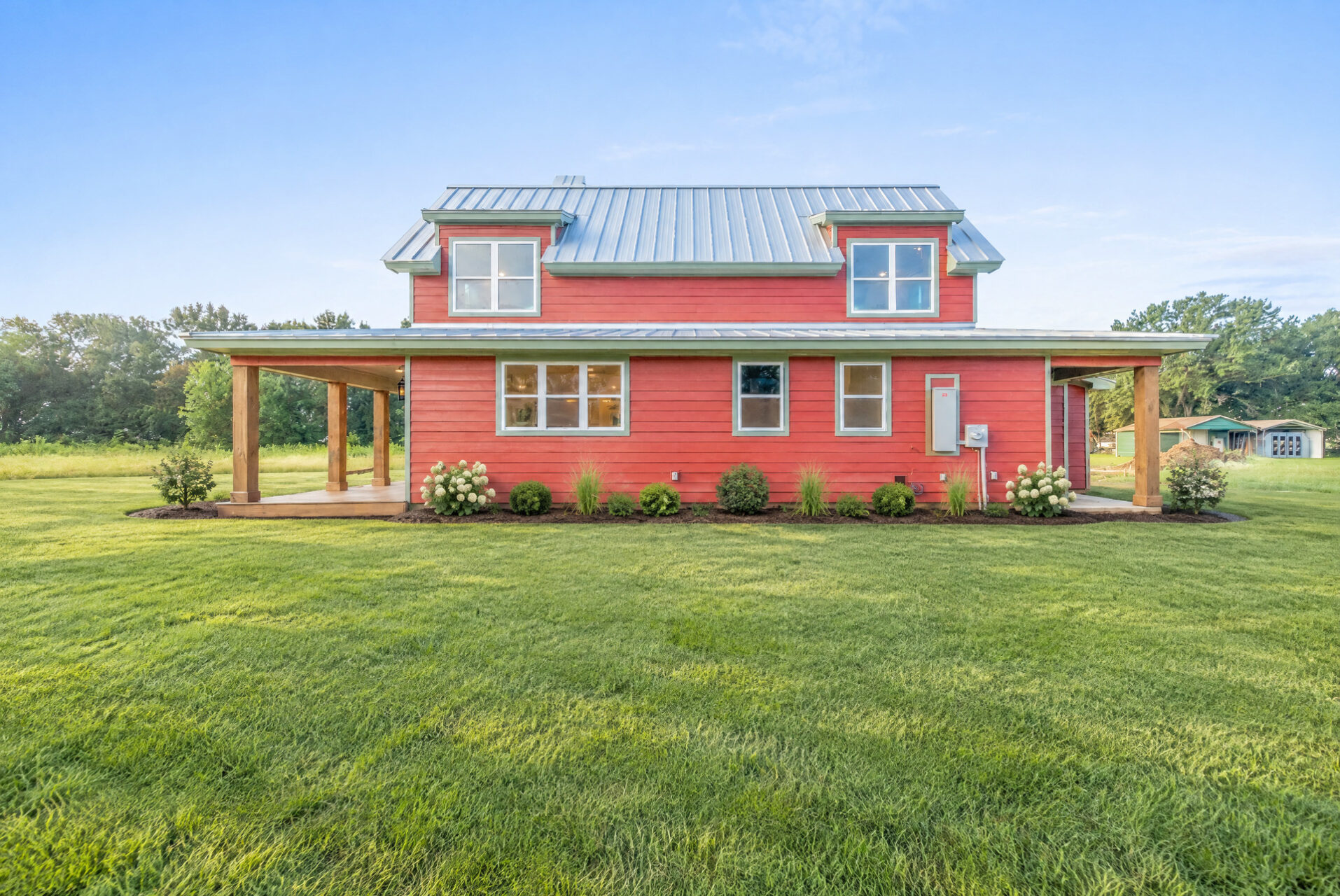 A two-story red farmhouse with a metal roof and a covered porch, set in a grassy field.