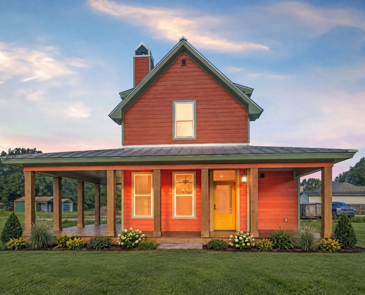 A two-story orange farmhouse with a wraparound porch and a bright yellow front door, set against a sunset sky.
