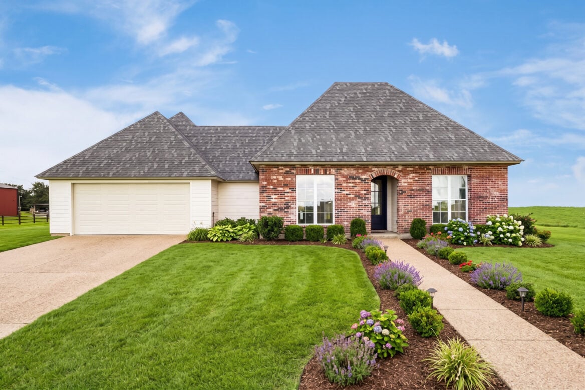 A single-story brick house with a two-car garage, a paved walkway, and front garden beds with flowering shrubs and perennials.